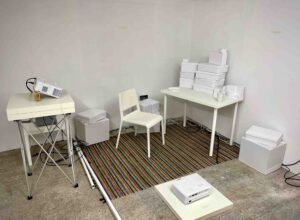 This image shows the set up I made for the installation described. It shows the corner of a gallery space, with white walls. There is a smallish, simple white table in the corner with various boxes, mugs and plates stacked up a bit haphazardly on it. It is a wide angle shot, showing how the projectors are arranged around this table. We can see a white chair near the desk, as if someone is about to sit down. On the left there is white plinth with a projector on it. There is another projector on the floor pointing at the back wall. The projectors are at 90 degrees to each other. You can’t see it on this image, but there is also a third projector mounted high up on the wall to the right of the main table, pointing downwards onto the table. There are some larger white boxes on the floor – each has a speaker inside it to help separate the soundtrack out spatially.