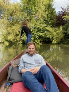 Photograph of Josh St Pierre sitting in a punt on the river in Oxford. He is pictured sitting centrally, smiling to camera, wearing a grey jumper and blue jeans. Behind him the punter, a woman in a black jumper and jeans, is looking backwards as she pushes against the river bed with a pole.