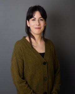 Photograph of Geneviève Lamoureux, a white woman with dark brown hair wearing a green cardigan, pictured smiling to camera and standing against a grey background 